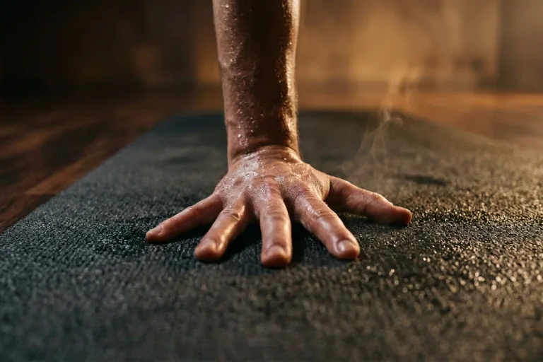 A sweaty bare hand pressed flat with fingers spread wide into a textured charcoal yoga mat, droplets on skin and mat and a thin steam wisp in warm key light inside an empty moody hot yoga room ? the best yoga mats for sweaty hands that improve grip through hot, humid sessions.